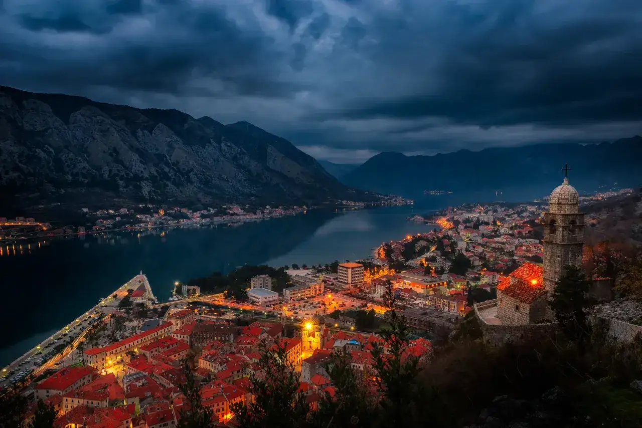 Wieczorny widok na zatokę i miasto Kotor w Czarnog&oacute;rze. Oświetlone budynki z czerwonymi dachami lśnią w blasku latarni, a g&oacute;ry tworzą mroczne tło.
