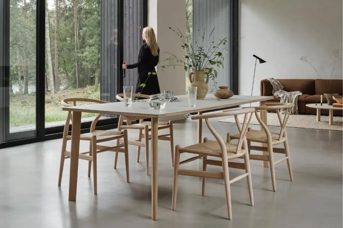 Ett modernt **stilleben matbord** med stolar i ljus tr&auml;, placerat vid stora f&ouml;nster med utsikt &ouml;ver skog. En kvinna st&aring;r vid f&ouml;nstret.