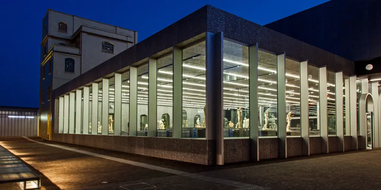Modern architecture houses classical sculptures in Milan art galleries, illuminated by bright lights at dusk.