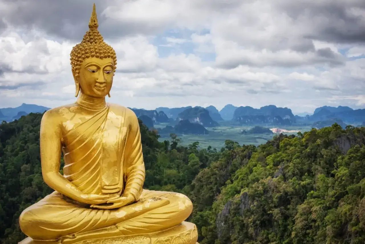Goldener Buddha thront &uuml;ber &uuml;ppiger Landschaft. Ein Weg f&uuml;hrt zu einem Tempel, vielleicht einer Tigerh&ouml;hle, mit vielen Stufen.