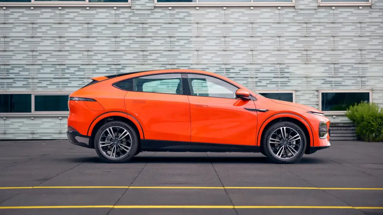 Un elegante coche eléctrico grande de color naranja brillante, con líneas deportivas y un diseño moderno, aparcado frente a un edificio con fachada texturizada.