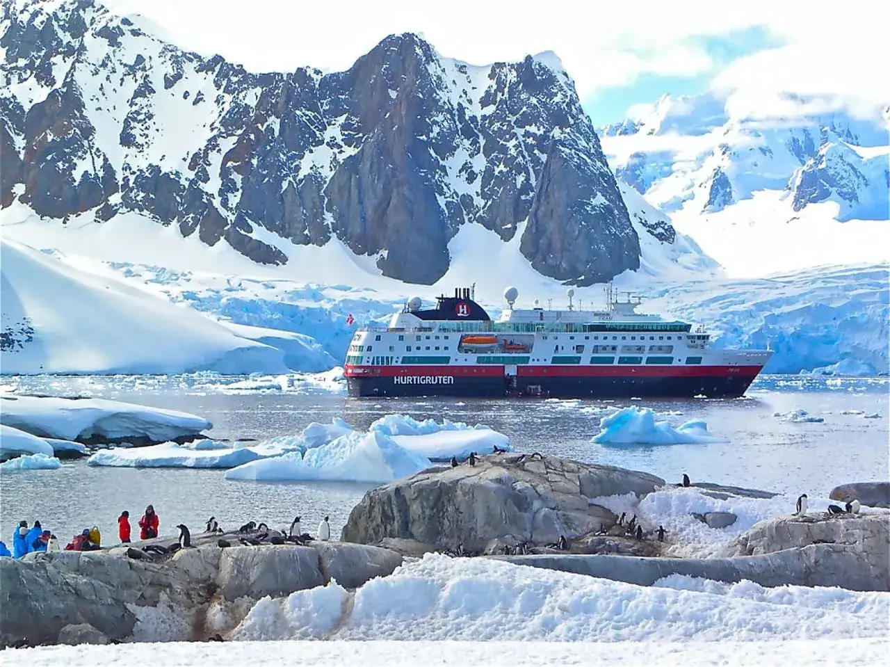 Statek wycieczkowy Hurtigruten na tle lodowych gór. Na pierwszym planie kolonia pingwinów i turyści. To krajobraz, który przypomina największą pustynię na świecie – Antarktydę.