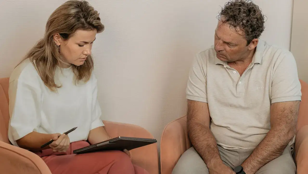 Een vrouw met een tablet en pen kijkt naar een man. Dit kan een beeld zijn van wanneer heeft relatietherapie geen zin, als er geen wil is om te veranderen.