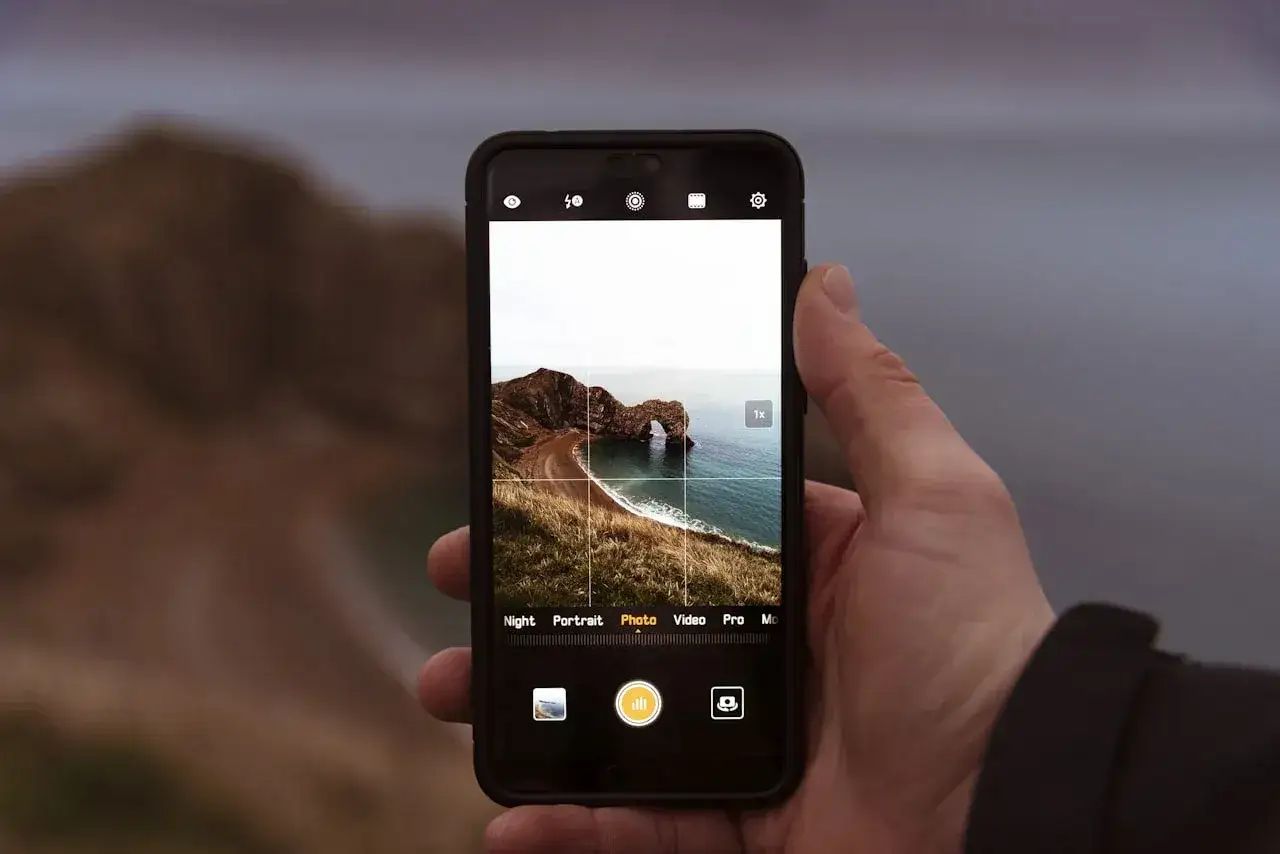 Dłoń trzyma smartfon z widokiem na skalny łuk Durdle Door. Aplikacja do wywoływania zdjęć jest otwarta, gotowa do uchwycenia piękna krajobrazu.