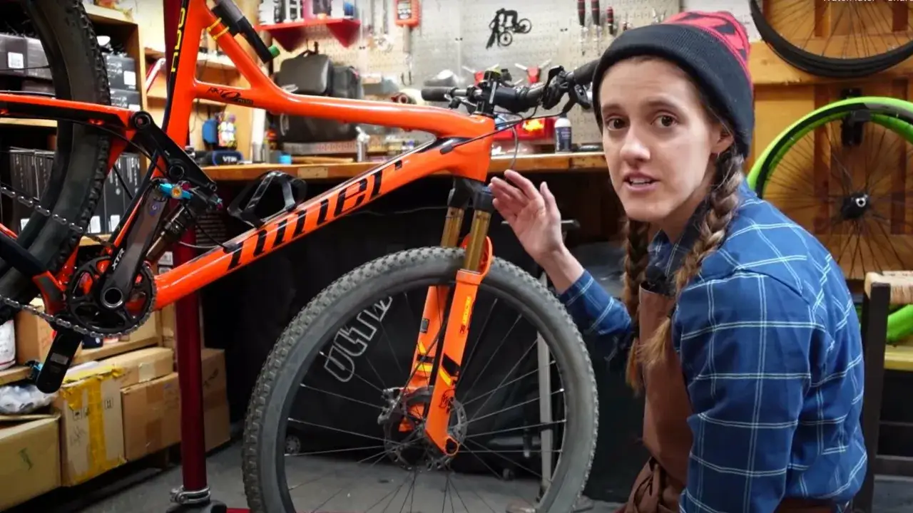 A woman in a plaid shirt and beanie explains bike maintenance next to an orange mountain bike on a stand.