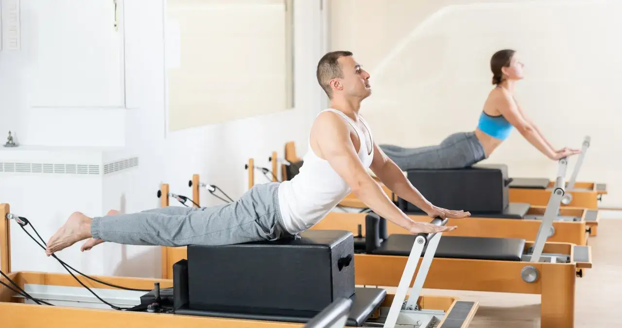 Mężczyzna i kobi ćwiczą na reformerach. To świetny spos&oacute;b, by dowiedzieć się, pilates co to jest i jak poprawić sylwetkę.