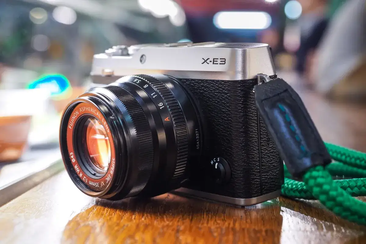 Elegancki aparat Fujifilm X-E3 z obiektywem Fujinon 35mm f/1.2 na drewnianym stole. Zielony pasek dodaje mu charakteru.