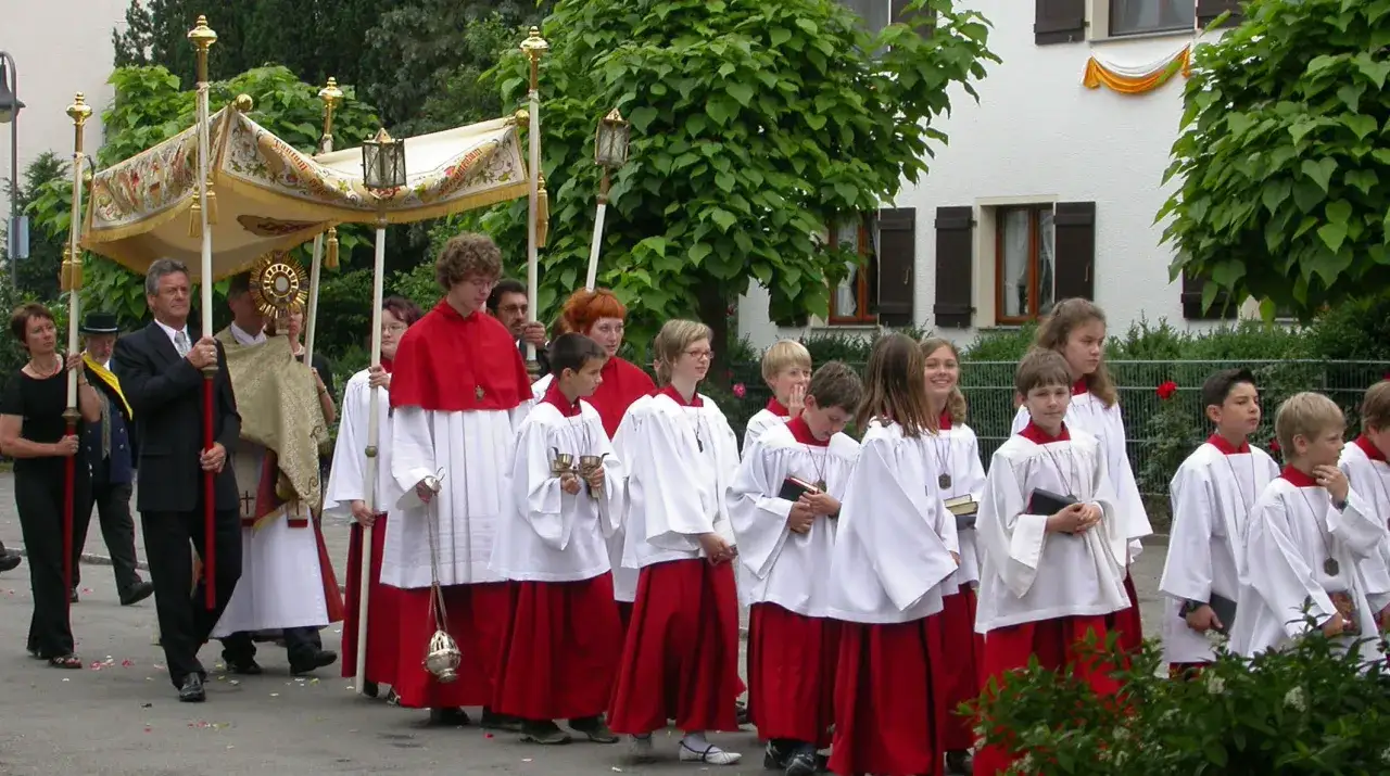 Prozession am Fronleichnam: Ministranten in weißen Alben und roten Röcken tragen eine Monstranz unter einem Baldachin.