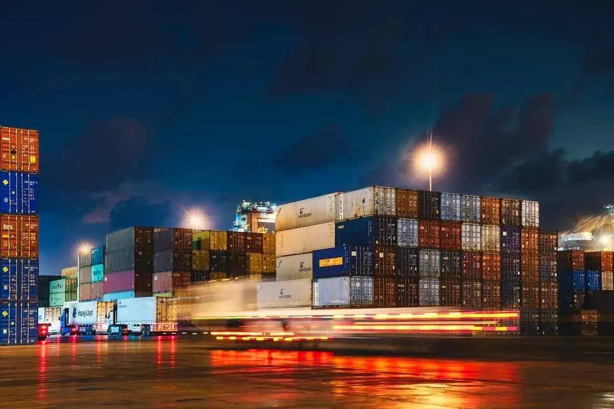 Nachts im Oman Port: Ein LKW fährt durch Reihen von Frachtcontainern, deren Lichter sich auf dem nassen Boden spiegeln.