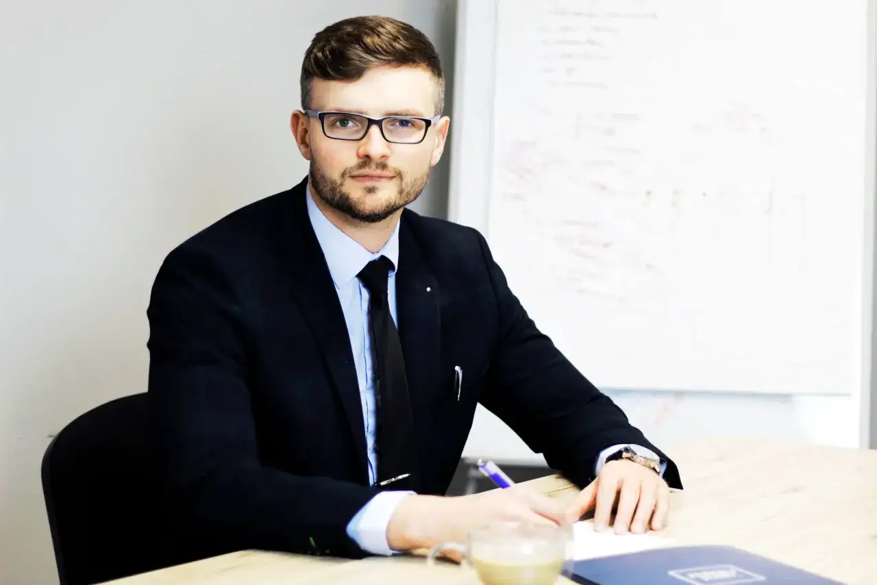 Mężczyzna w garniturze i okularach siedzi przy biurku, pisząc coś na kartce.