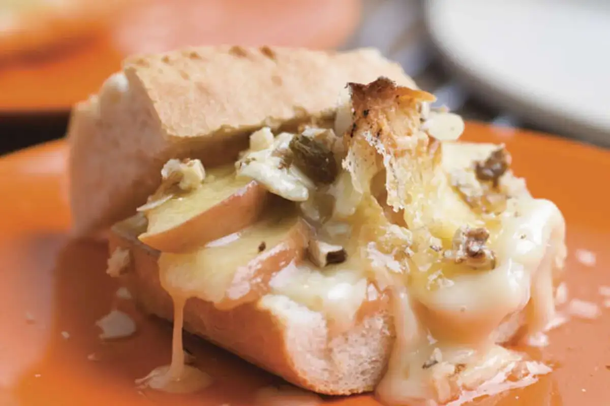 Un camembert fondant, préparé à l'air fryer, déborde sur une tranche de pain avec des morceaux de pomme et des noix.
