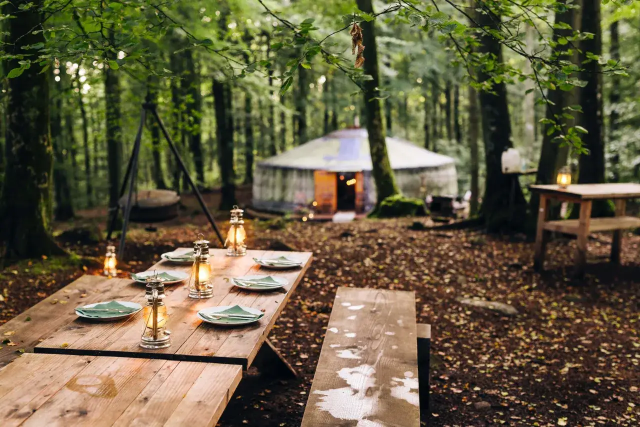 En uppdukad middag med lyktor och tallrikar väntar vid ett träbord i skogen. I bakgrunden syns en yurt, en inbjudande plats för glamping i Skåne.