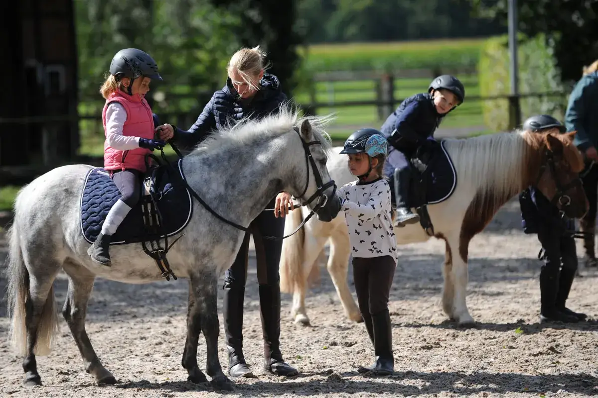 Kinder reiten auf Ponys, eine Frau hilft einem M&auml;dchen auf dem grauen Pony.