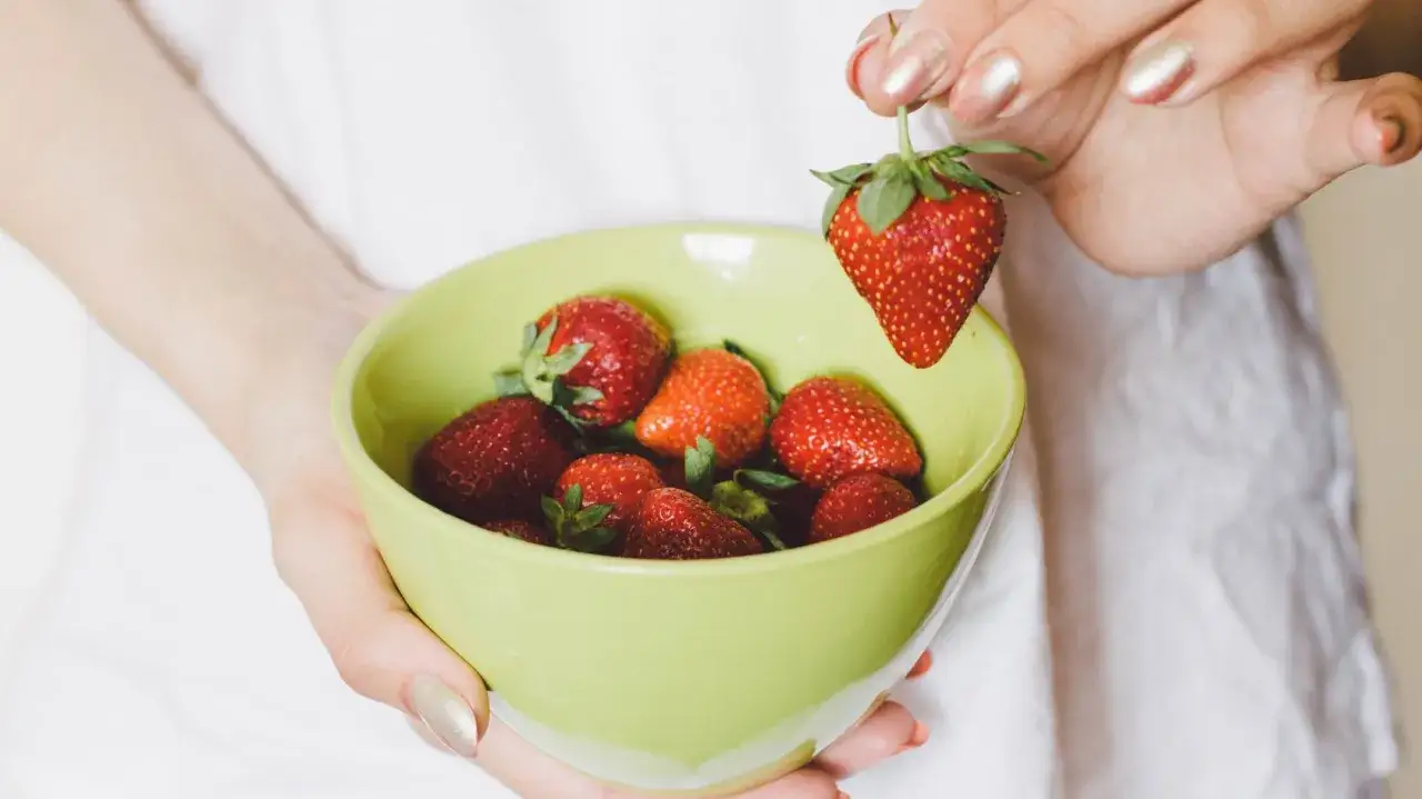 Hände halten eine hellgrüne Schüssel mit frischen Erdbeeren. Eine einzelne Erdbeere wird über der Schüssel gehalten, bereit zum Waschen.