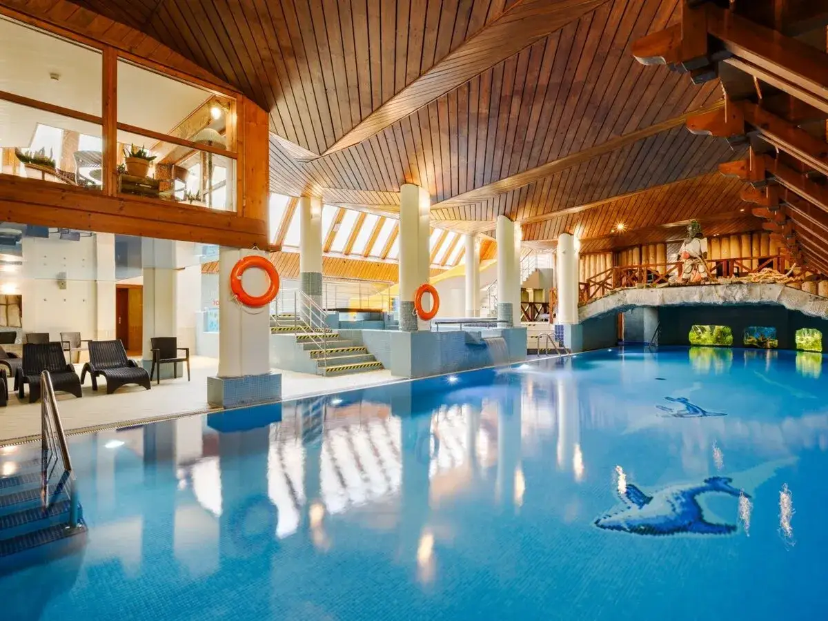 Basen w hotelu Belvedere z drewnianym sufitem i mostkiem. Woda jest błękitna, a na dnie widać mozaiki z ptakami.