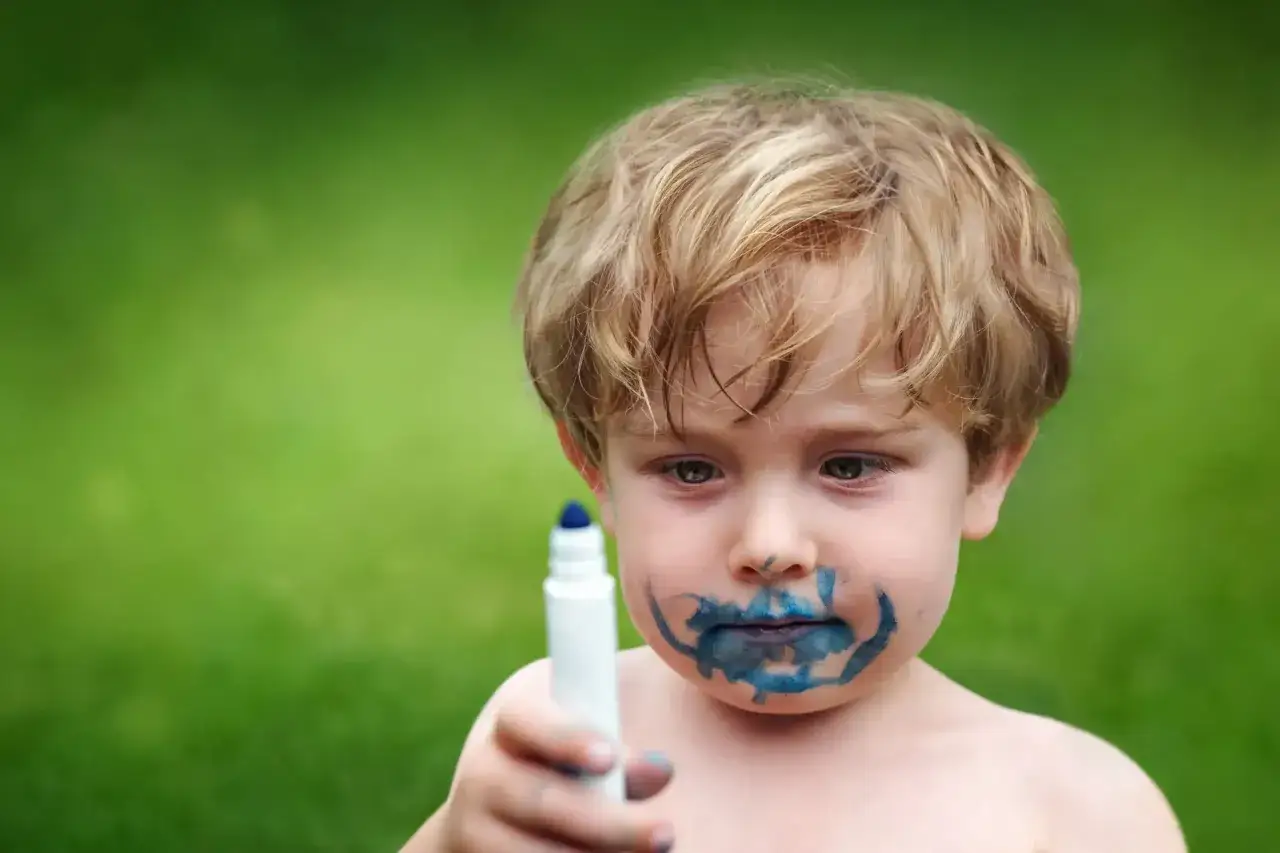 Kleiner Junge mit blauem Filzstift im Gesicht, der versucht, ihn von der Haut zu entfernen.