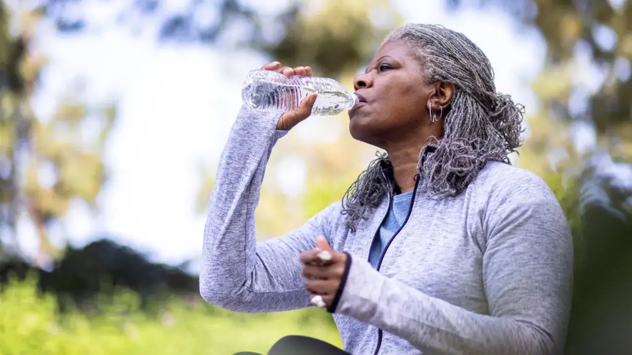 Femme adulte aux cheveux gris buvant de l'eau d'une bouteille. Elle porte une veste grise. L'eau cristalline est essentielle pour &eacute;viter la diarrh&eacute;e chez l'adulte.