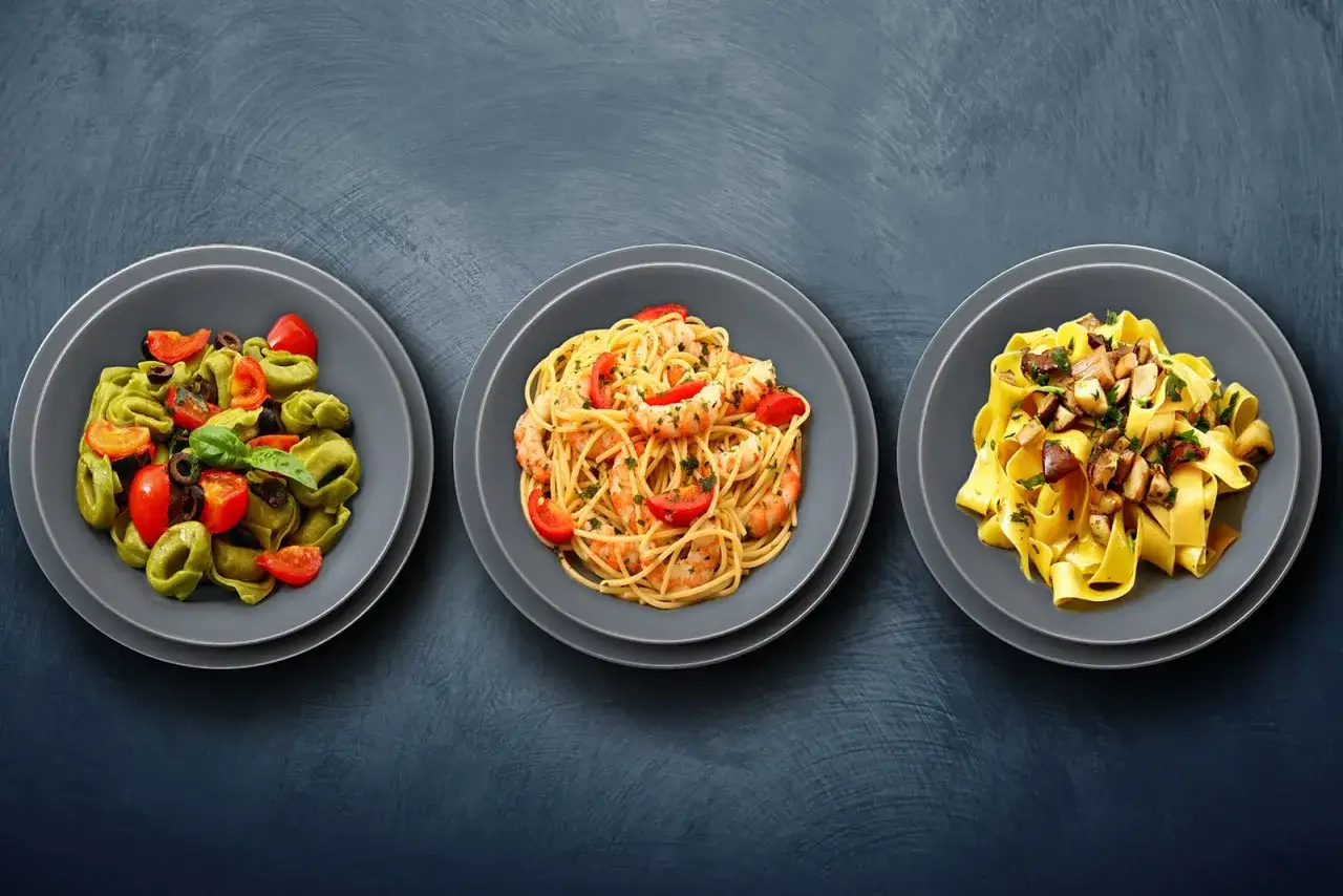 Trzy talerze z różnymi rodzajami makaronu: zielone tortellini z pomidorami i oliwkami, spaghetti z krewetkami i pomidorkami, oraz tagliatelle z grzybami.
