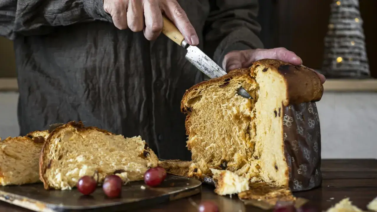 Man corta un panettone con un cuchillo. Así se come el panettone, con uvas rojas al lado.