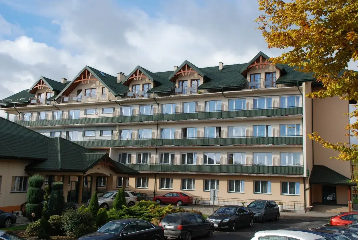 Sanatorium Plon Polańczyk z zielonym dachem i balkonami, otoczone drzewami. Przed budynkiem zaparkowane samochody.