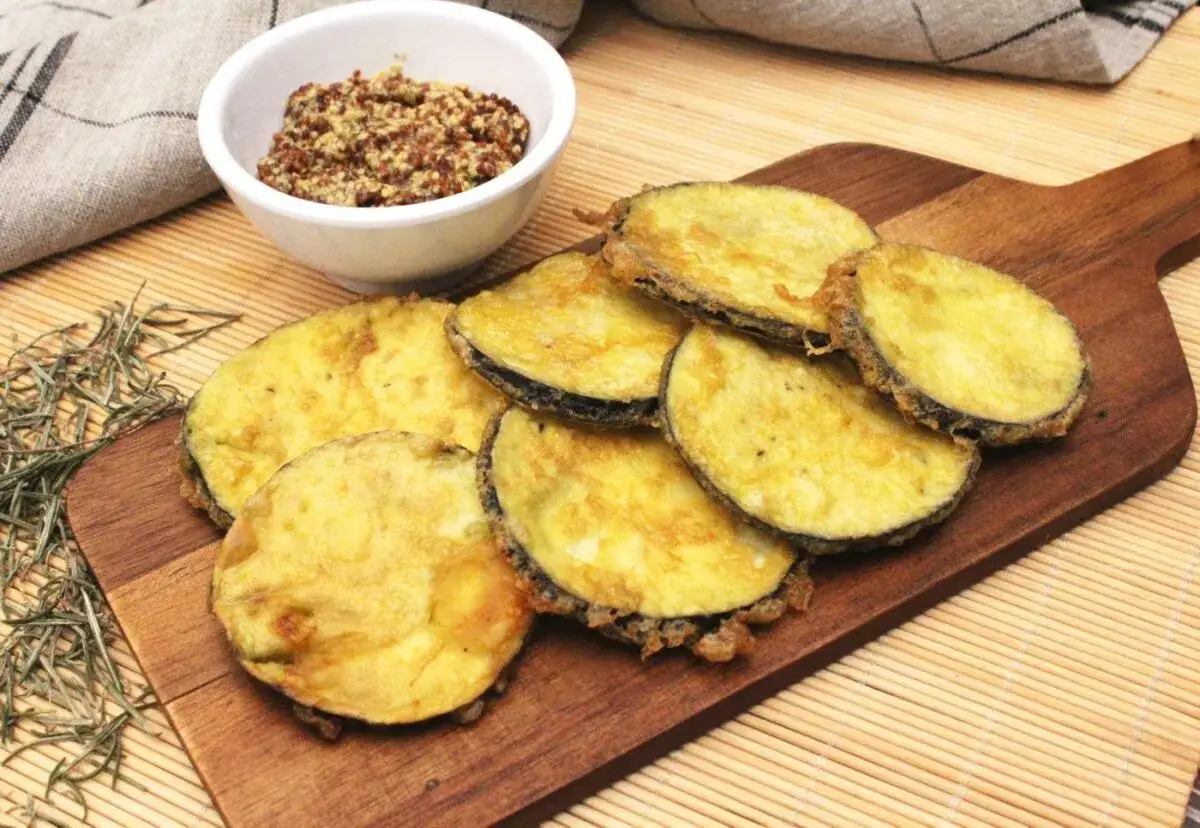 Berenjenas rebozadas doradas y crujientes, servidas en tabla de madera con un cuenco de mostaza de grano entero.