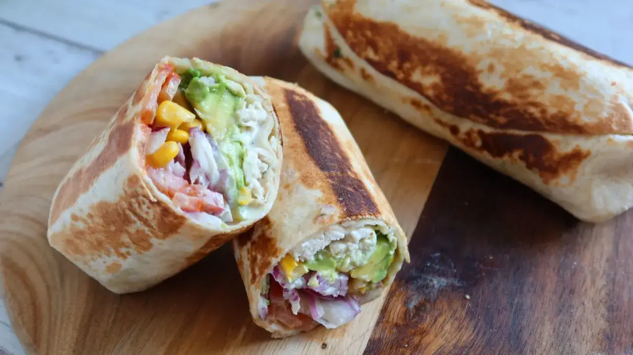 Burritos de pollo recién hechos, rellenos de aguacate, maíz, tomate y cebolla morada, servidos en una tabla de madera.