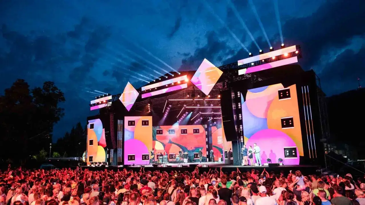 Koncert na żywo podczas wakacyjnej trasy dwójki. Tłum ludzi pod sceną, artyści na scenie, kolorowe ekrany LED i światła.