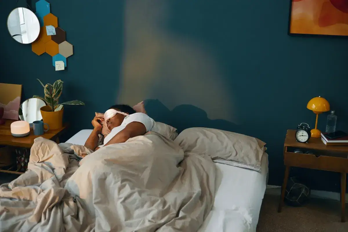 Osoba śpi w ł&oacute;żku z zasłoną na oczach. W sypialni dominują uspokajające kolory do sypialni, takie jak granatowa ściana i beżowa pościel.