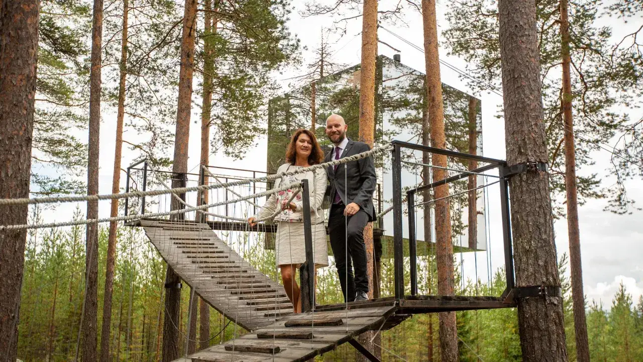 Ett par står på en hängbro framför ett spegelhus i skogen i Harads, Sverige.