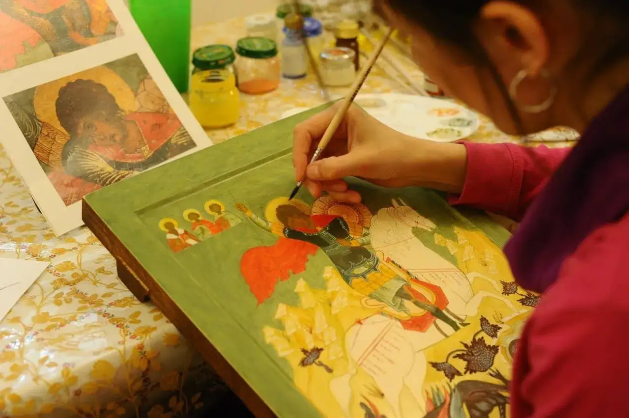 Una persona pinta con temperas un icono religioso. Se ve una mano con un pincel, detalles de la pintura y frascos de pintura.