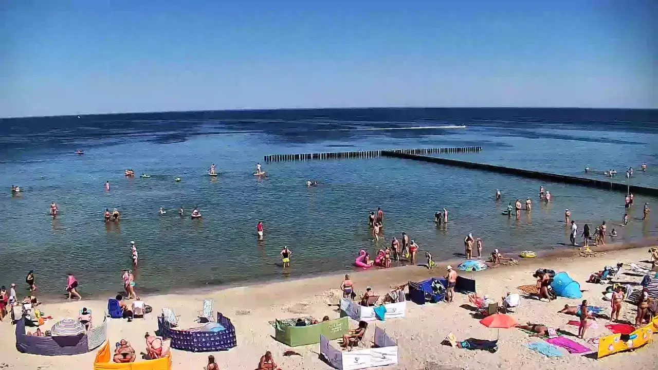 Słoneczny dzień nad morzem, ludzie kąpią się i opalają na plaży. W oddali widać falochron i łódź. Dźwirzyno molo tętni życiem.