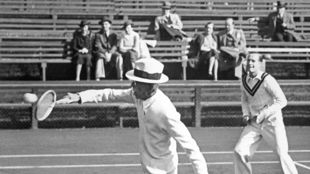 Gustaf V i tennisdr&auml;kt sl&aring;r en boll p&aring; en grusplan, publiken sitter p&aring; l&auml;ktaren.