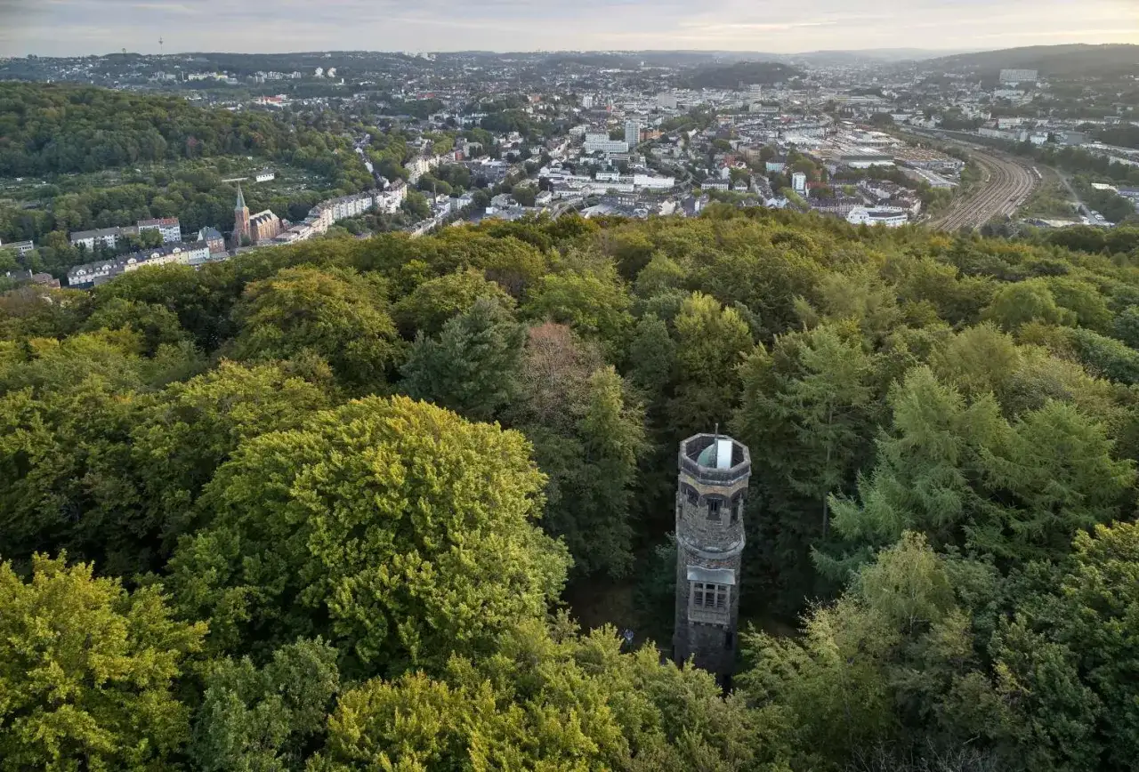 Der von der Heydt Turm ragt aus einem dichten gr&uuml;nen Wald hervor, mit einer Stadt im Hintergrund.