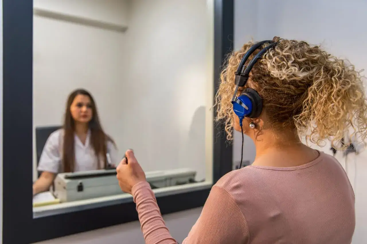 Frau mit Kopfhörern im Audiometrie-Raum, um Tinnitus festzustellen. Eine Ärztin im Hintergrund beobachtet.
