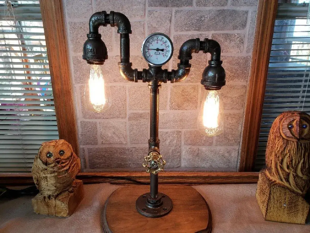 Oryginalna lampa z rurek w stylu industrialnym, z dwoma żarówkami i manometrem. Obok stoją rzeźbione sowy.