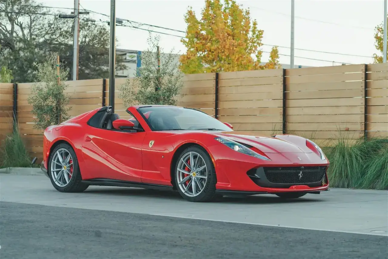 Czerwony kabriolet Ferrari 812 GTS z opuszczonym dachem, zaparkowany przed drewnianym płotem i zielenią.