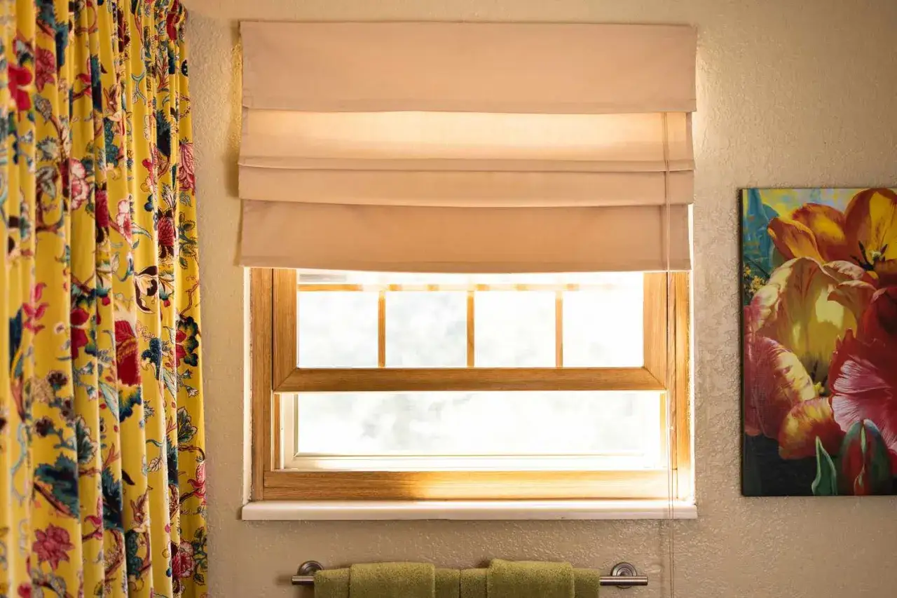 Learn how to make roman shades for a stylish window treatment. This image shows a window with a beige roman shade, a floral yellow curtain, and a tulip painting.