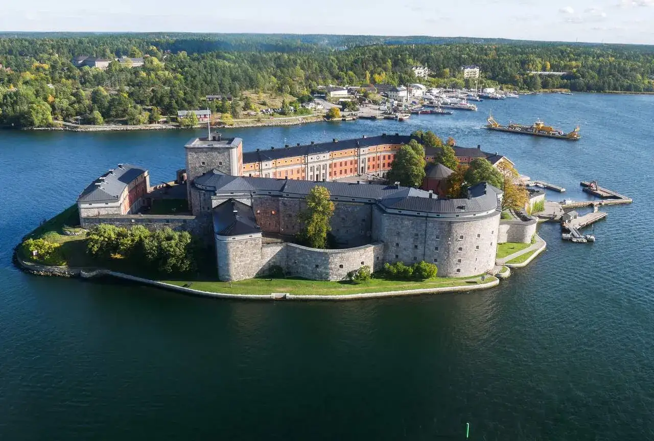 Vaxholms kastell, en imponerande fästning på en ö, omgiven av vatten och grönskande skog. En gul färja syns i vattnet.