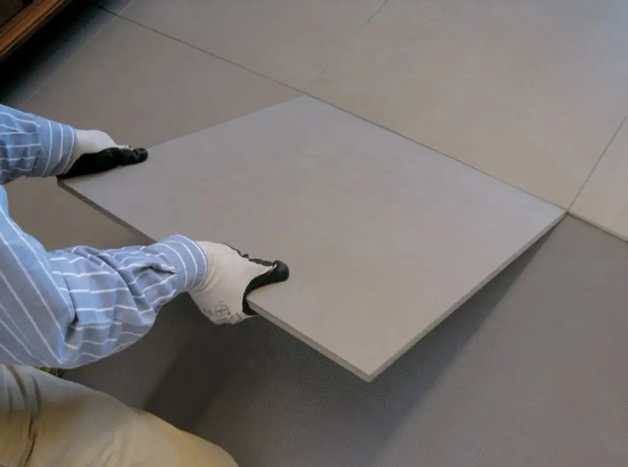 Pose d'une nouvelle dalle de carrelage sur carrelage existant. Mains gant&eacute;es tenant la nouvelle pi&egrave;ce.