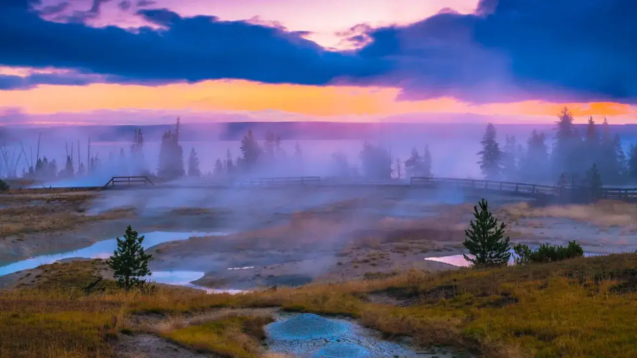 Mglisty poranek w **parku narodowym Yellowstone**. Drewniana kładka wije się wśród parujących źródeł i turkusowych kałuż, a nad wszystkim unosi się fioletowo-pomarańczowe niebo.