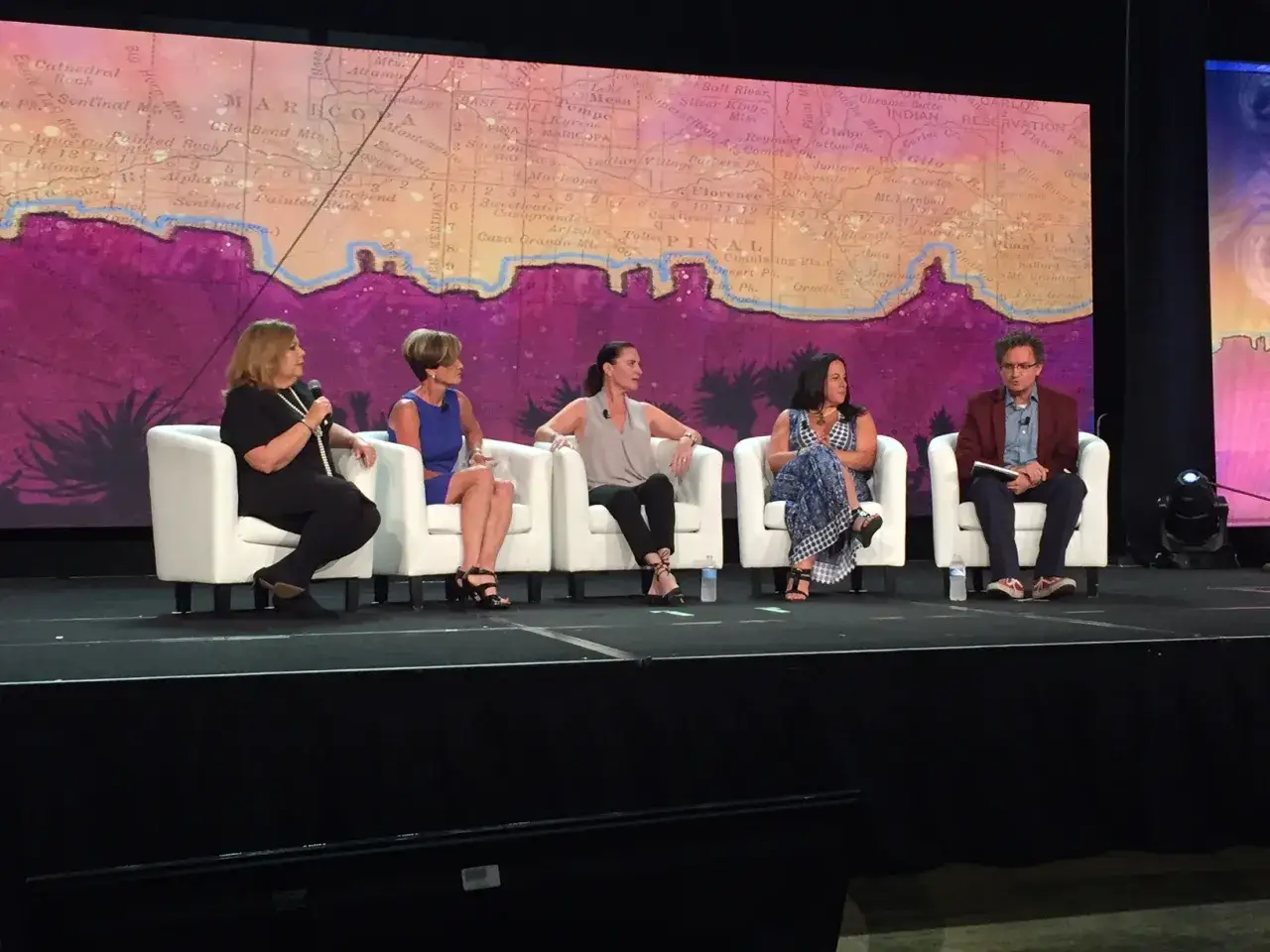 Five people sit on white chairs on a stage, discussing how to moderate a panel. A large map is projected behind them.