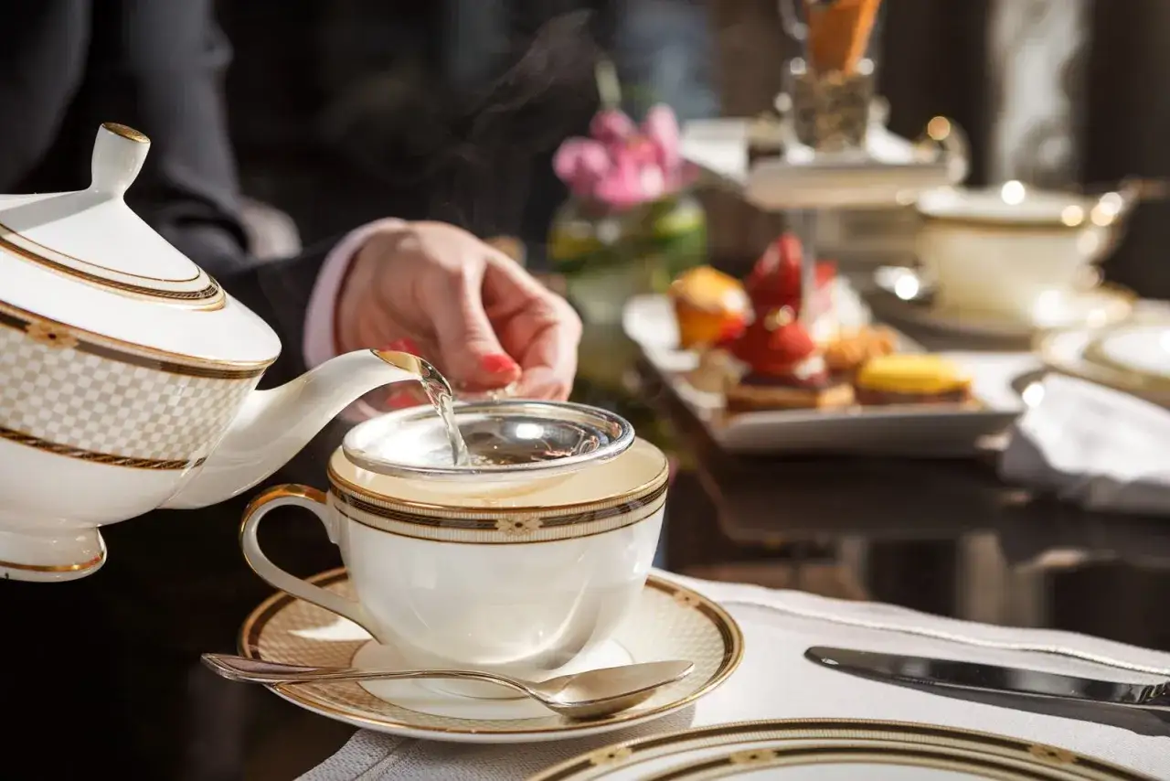 En person häller upp te från en elegant tekanna i en kopp, redo för en klassisk afternoon tea med utsökta bakverk i bakgrunden.