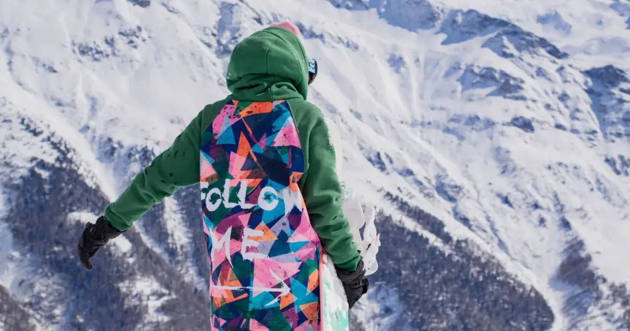 Osoba w kolorowym, wzorzystym ubiórze na narty, z deską snowboardową, stoi na tle ośnieżonych gór.