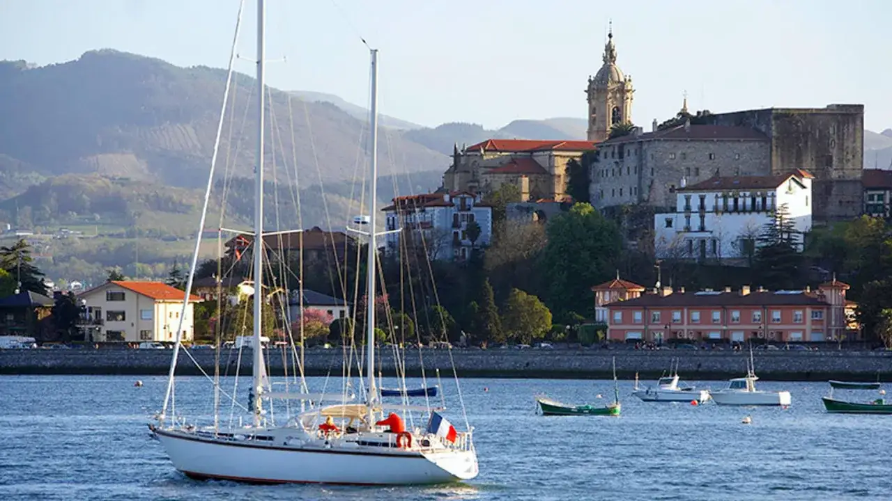 Velero navega en la bahía de Hondarribi, con el casco antiguo y montañas de fondo.