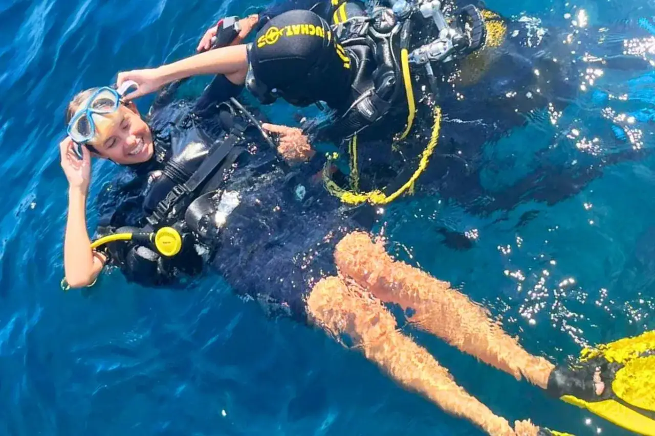 Un jeune plongeur souriant, en équipement complet, est aidé par un moniteur lors d'une colonie de vacances plongée. L'eau bleue scintille autour d'eux.