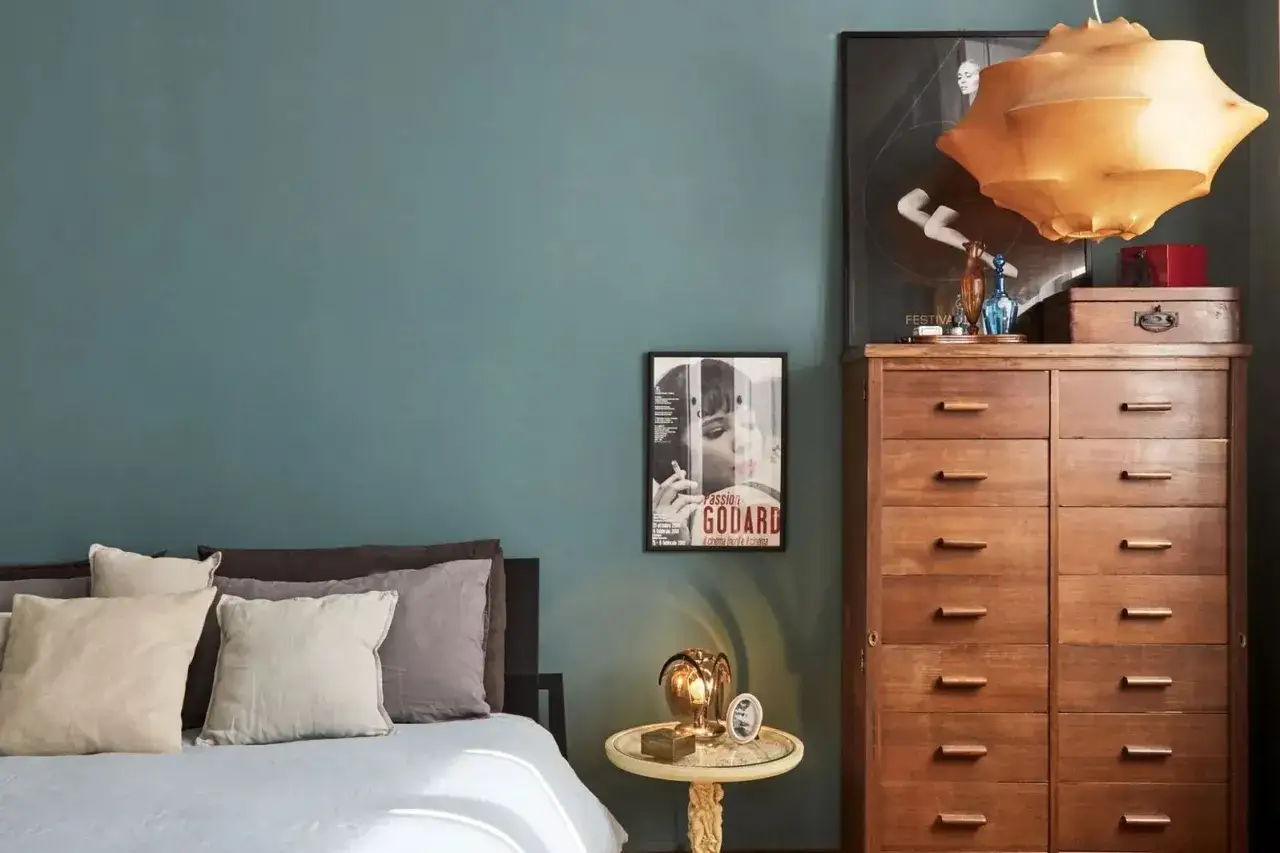 Camera da letto con pareti color ottanio, un comò in legno scuro e un letto con cuscini in tonalità neutre. L'abbinamento colori crea un'atmosfera rilassante.
