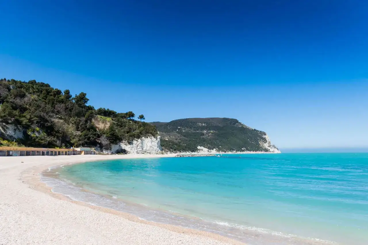 Spiaggia bianca e mare turchese nelle Marche, ideale per vacanze al mare. Scogliere verdi e cielo azzurro completano il paesaggio.