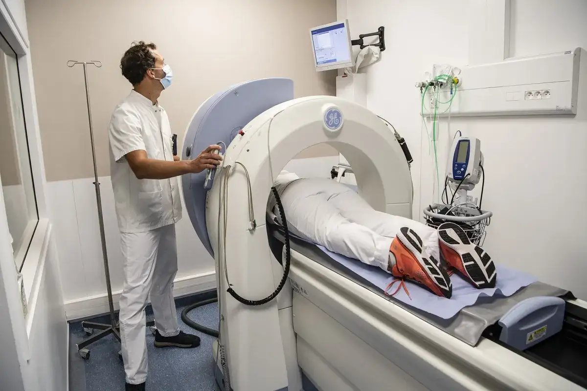 Un patient est allongé sur une table d'examen pour un PET scan, un examen d'imagerie médicale. Un technicien en blouse blanche surveille l'appareil.