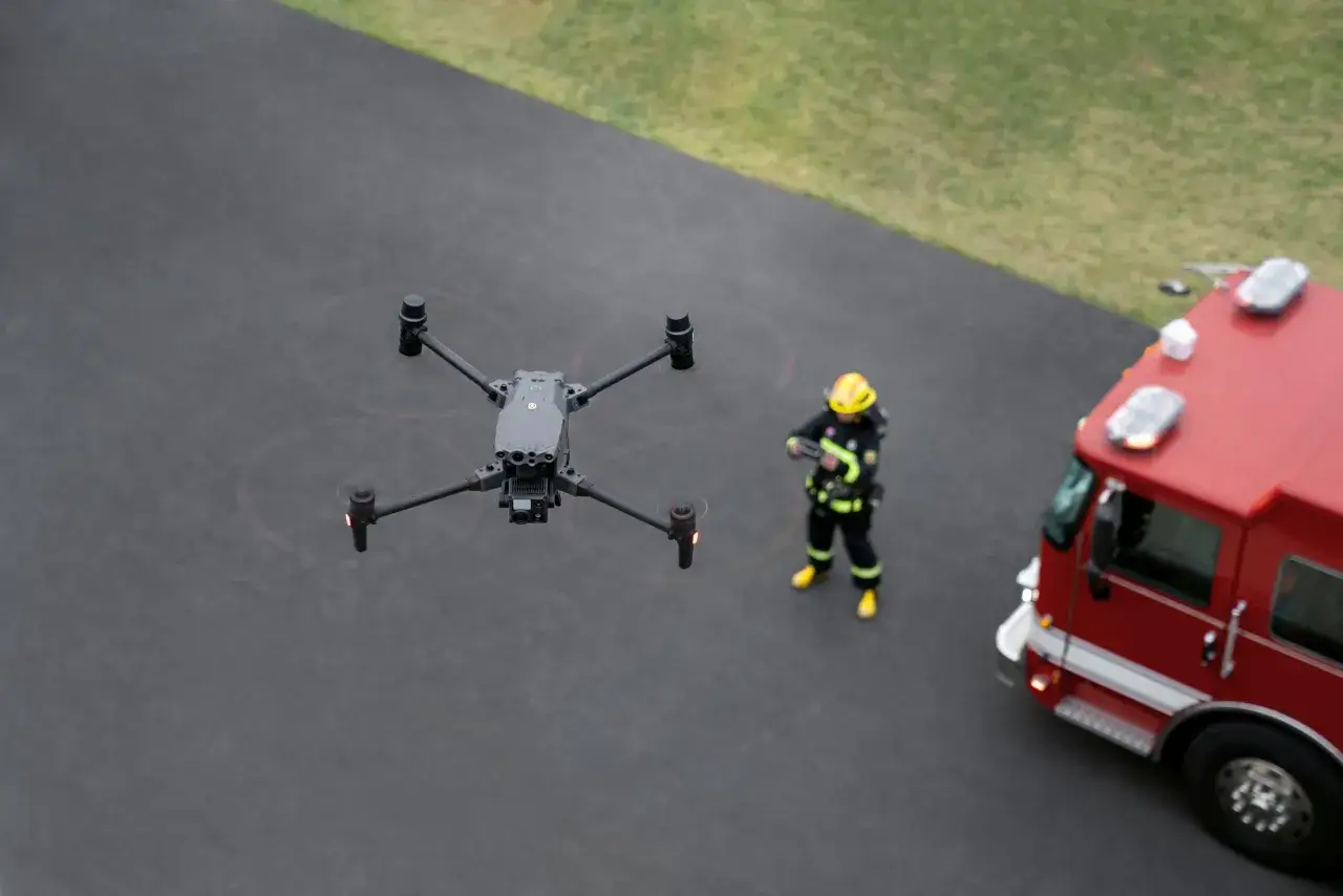 Strażak w żółtym hełmie i czarnym mundurze obserwuje drona, który unosi się nad asfaltem obok wozu strażackiego. Nowoczesne technologie w akcji KSRG.