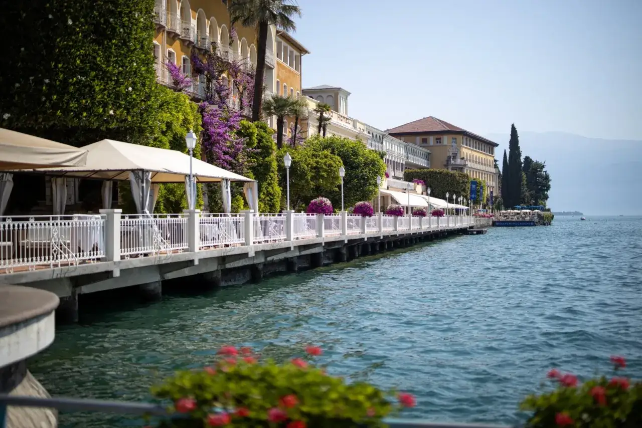 Un elegante lungolago a Gardone Riviera con hotel storici, pergolati fioriti e il blu del lago.